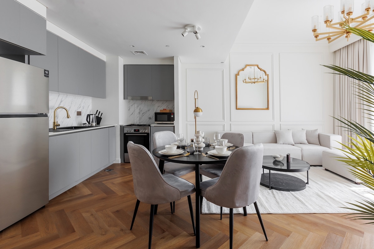 A modern kitchen is visible, featuring sleek gray cabinetry and countertops. A dining table, surrounded by upholstered chairs, is set for four. A comfortable living area is seen with a sectional sofa and a decorative coffee table. Natural light enters through a large window, enhancing the spacious layout.