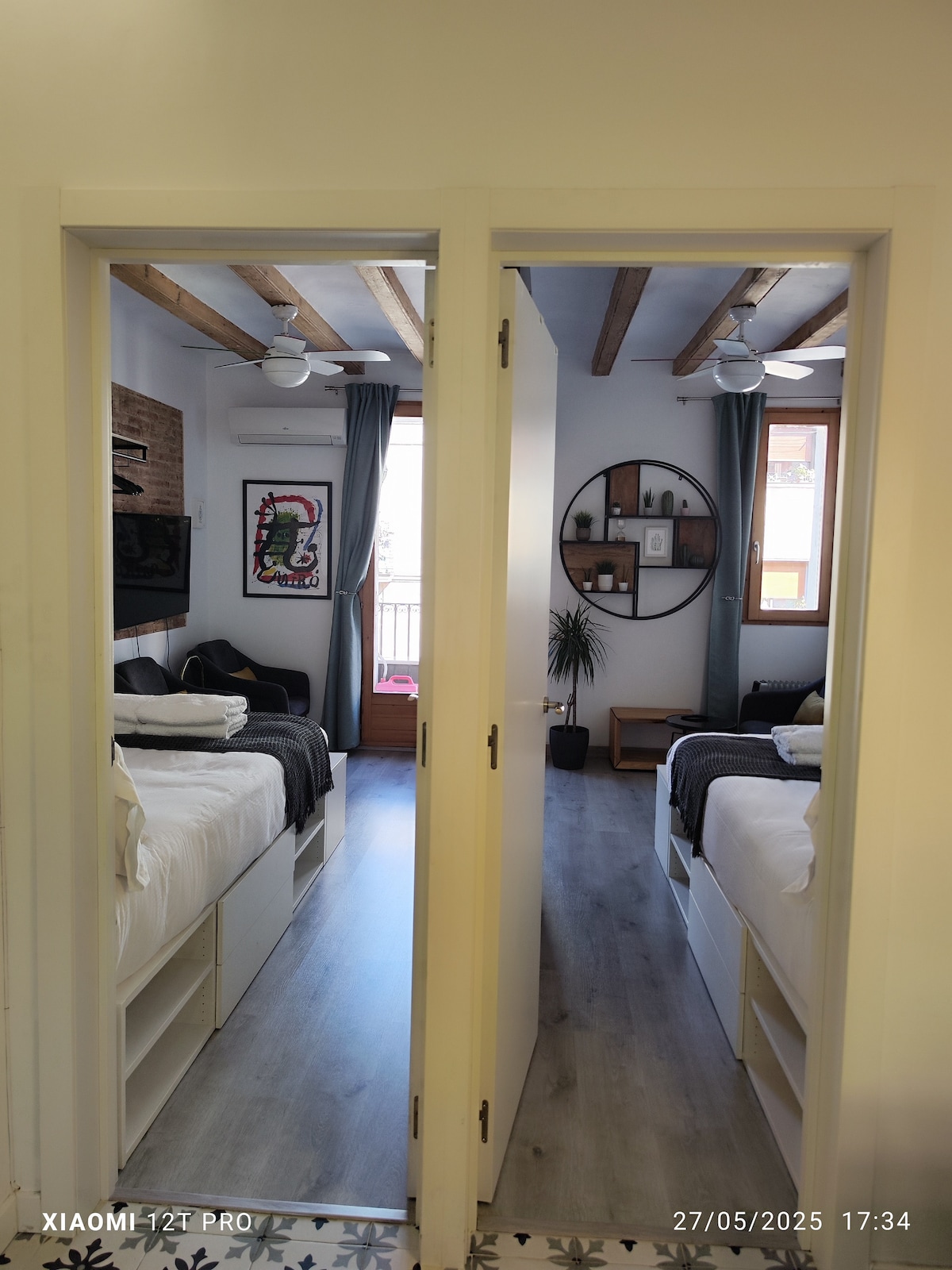 Two bedrooms are viewed through a double doorway, featuring modern design elements. Each room is furnished with a spacious bed and adorned with light decor. Natural light enters from windows, and items like artwork and plants are observed, adding character to the space.