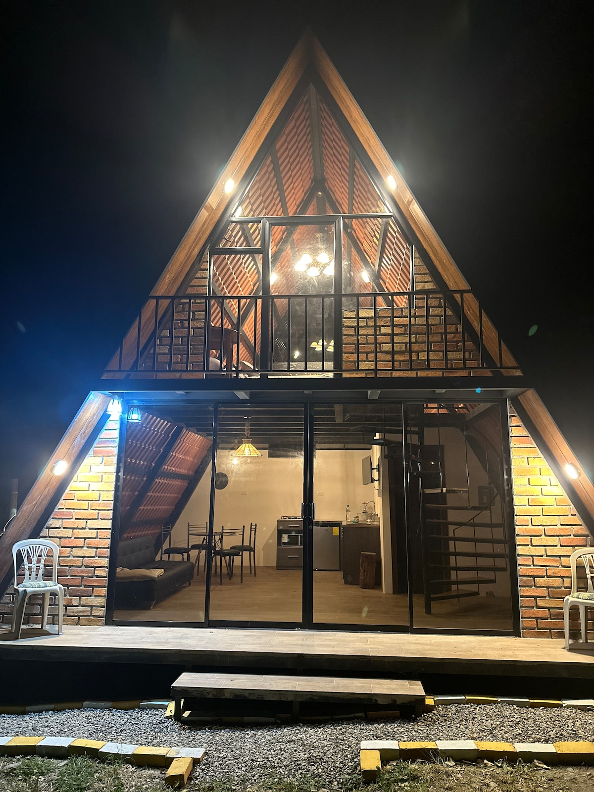 The unique triangular structure is illuminated at night, showcasing its warm brick facade and large glass windows. Two white chairs are positioned on the porch, inviting relaxation, while the interior is visible, highlighting the open living space and cozy atmosphere.