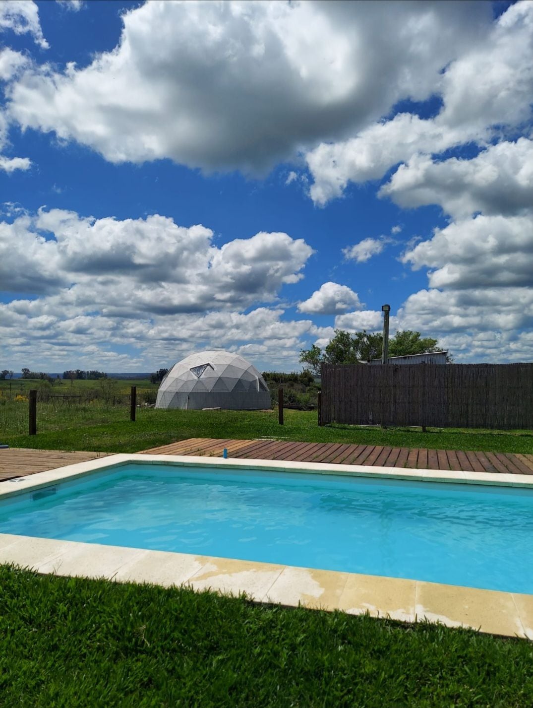 A shared swimming pool is set against a backdrop of lush green fields and a geodesic dome in the distance. The clear blue sky is dotted with fluffy white clouds, enhancing the outdoor landscape. Wooden decking surrounds the pool, providing a relaxing space for guests.