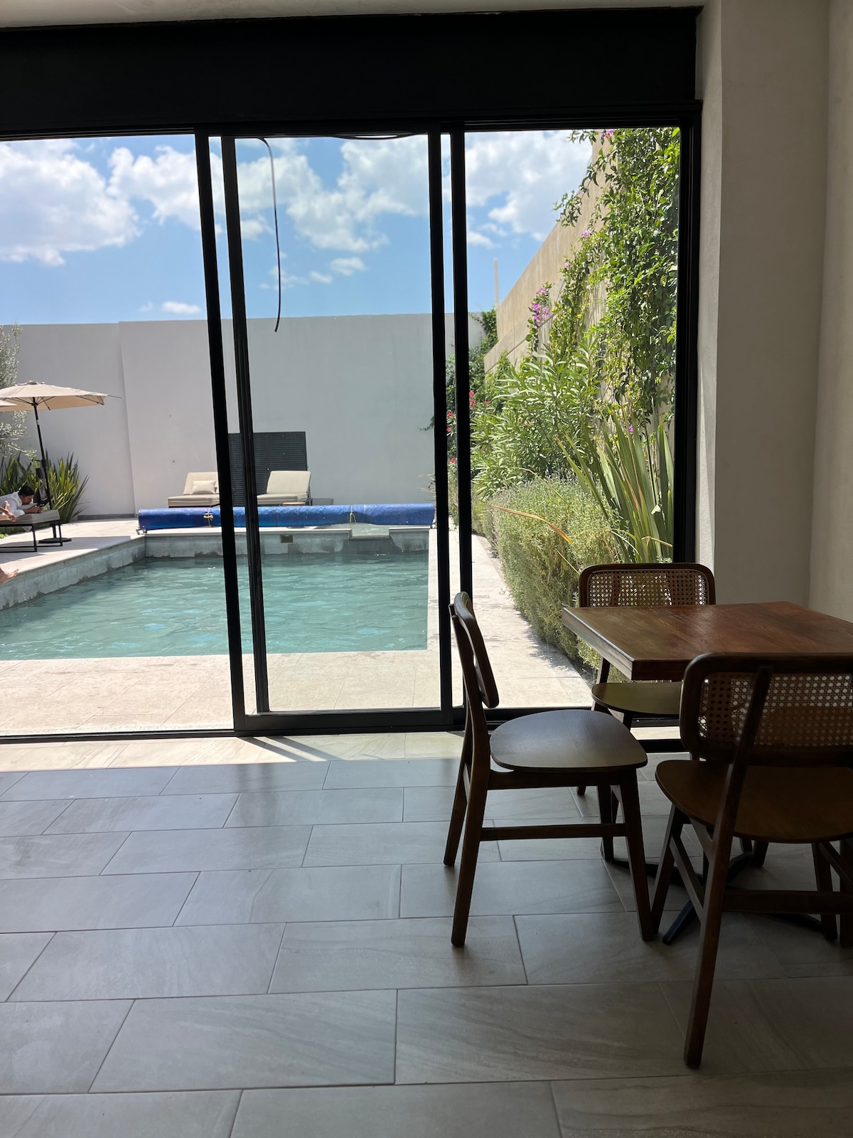 A dining area features a wooden table with two woven chairs, positioned near expansive glass doors. The doors lead to an inviting pool area, framed by lush greenery and outdoor lounging space, allowing natural light to fill the interior.