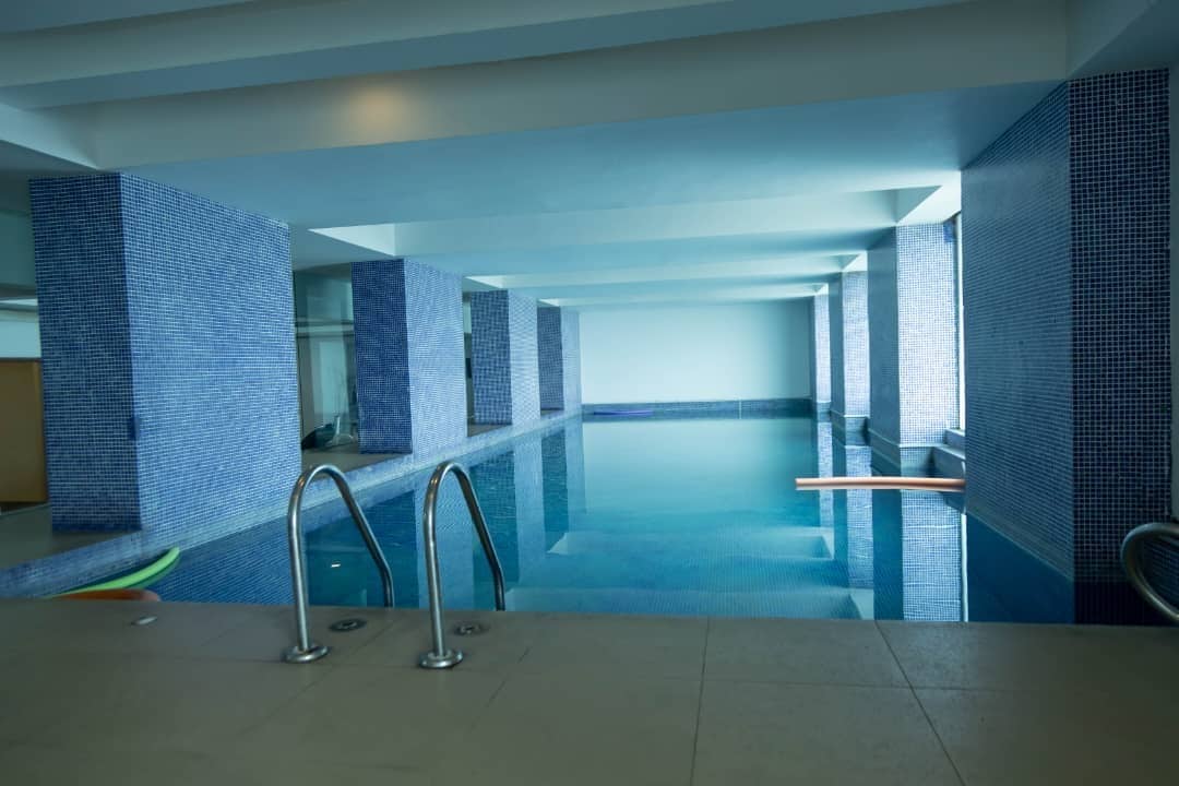 An indoor swimming pool features tranquil blue tiles lining the walls and floor. Steps lead into the water, and the area is illuminated by soft lighting. Colorful pool floats rest against the edge, creating a calm and inviting atmosphere for leisure activities.