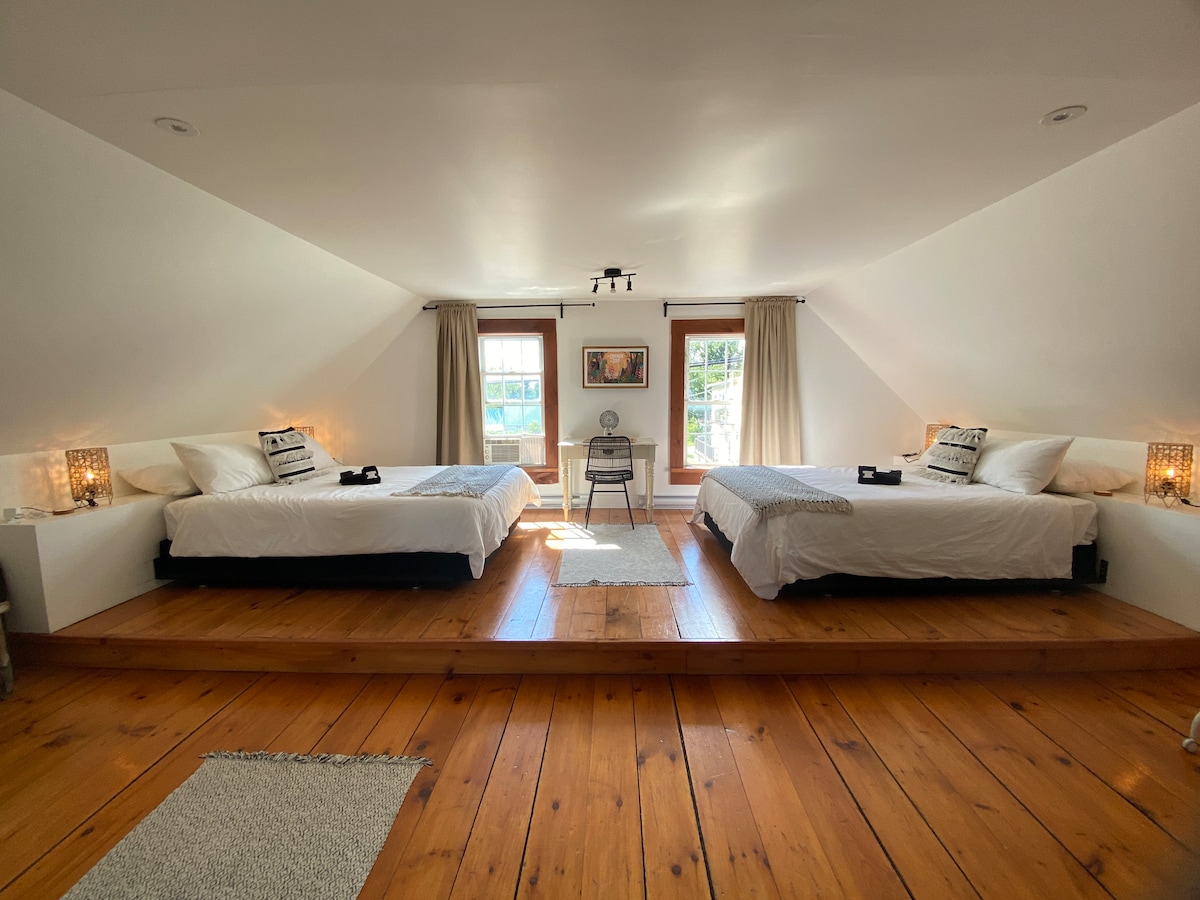 This spacious bedroom features two Queen beds positioned on a raised wooden platform, complemented by light-colored bedding. Natural light filters in through large windows adorned with sheer curtains, while a small table and decor items enhance the inviting ambiance. A textured area rug adds warmth to the wooden floor.
