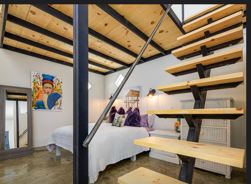 An open loft area displays a spacious bed adorned with decorative pillows. A vibrant piece of artwork hangs above the bed, complementing a full-length mirror nearby. Distinctive wooden stairs lead to the upper level, contrasting with the smooth concrete floor.