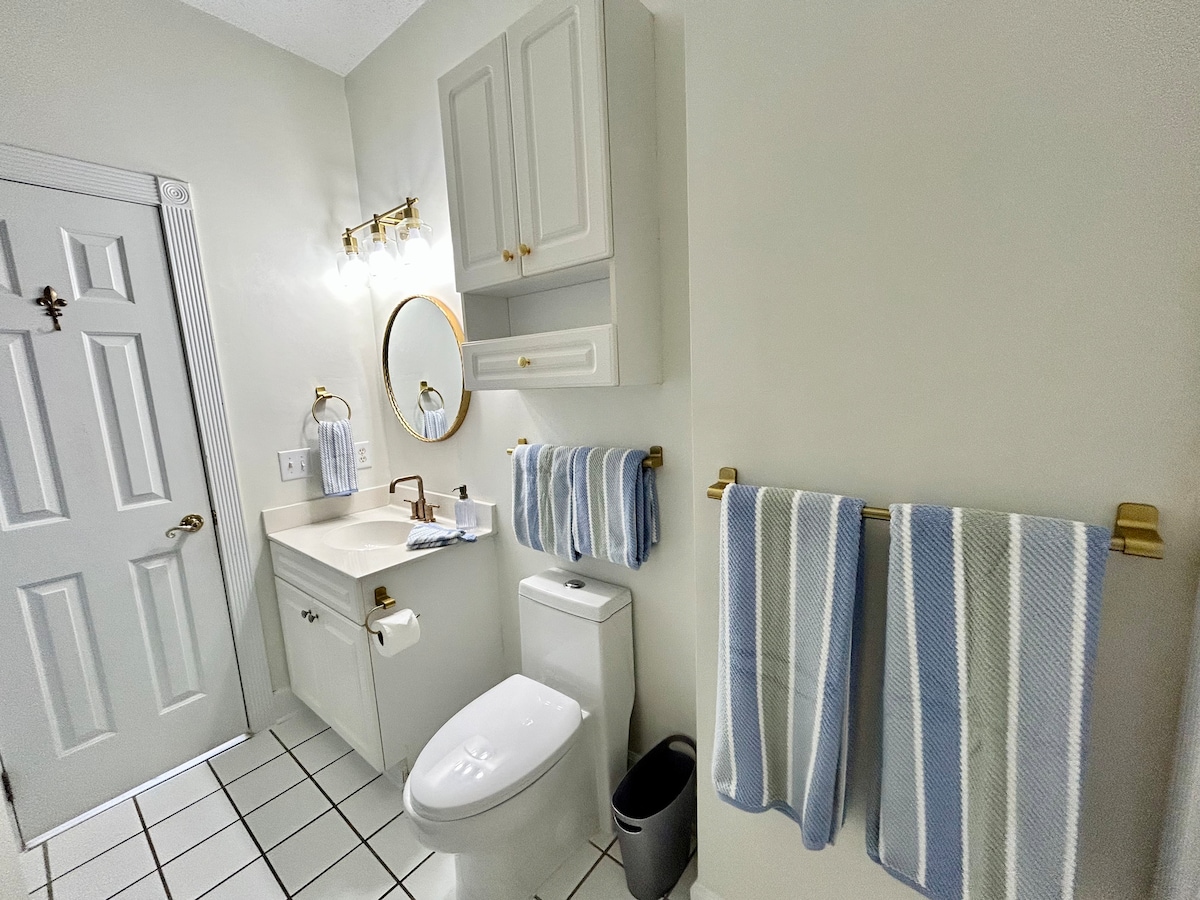 The bathroom features white cabinetry and a light-colored countertop, with a round mirror above the sink. Soft blue and white striped towels hang beside the toilet, complemented by a small waste bin. Ample natural light brightens the space through a window.