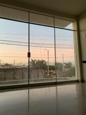A large glass door fills the space with natural light, offering a view of the outdoors. The floor is finished with light tiles, complementing the serene color palette. Palm trees and power lines are visible beyond the clear glass, framed by a soft pastel sky.