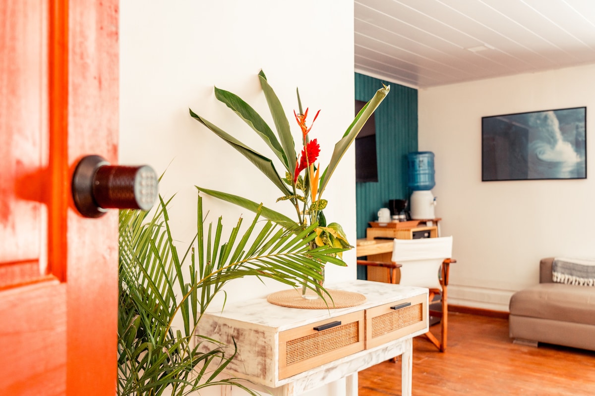 A welcoming entry area features a decorative table adorned with a tropical plant and vibrant flowers. The wooden floor complements the light-colored walls, while a water cooler is visible in the background, alongside a cozy seating area, providing an inviting space upon entering.