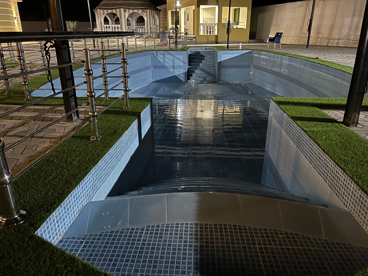 A uniquely designed pool area features varying depths, bordered by neatly trimmed grass. The pool's tiles reflect light at night, highlighting the structure's contours and steps. Decorative railings provide safety along the edges, blending functionality with a sleek aesthetic.