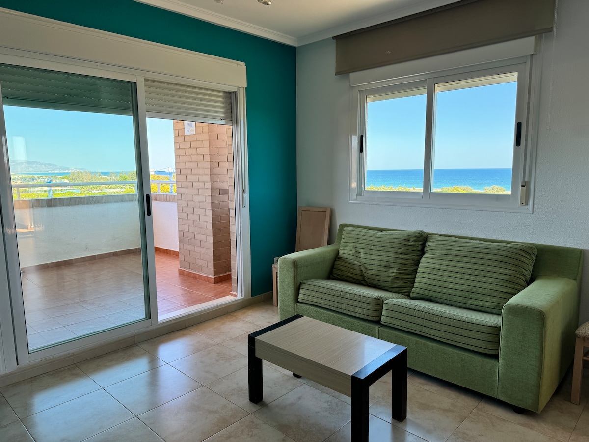 A spacious living area is highlighted by a green sofa and a small coffee table. Large windows allow natural light to fill the space, opening to a picturesque balcony with views of the sea and surrounding landscape. The walls are painted in a calming teal.