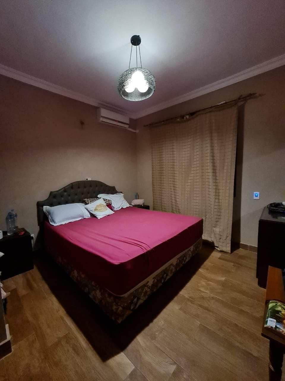 A cozy bedroom features a spacious bed dressed in a vibrant pink coverlet. Soft lighting is provided by a pendant light overhead, complementing the warm wall tones. A pair of bedside tables flank the bed, and an air conditioning unit is mounted on the wall.