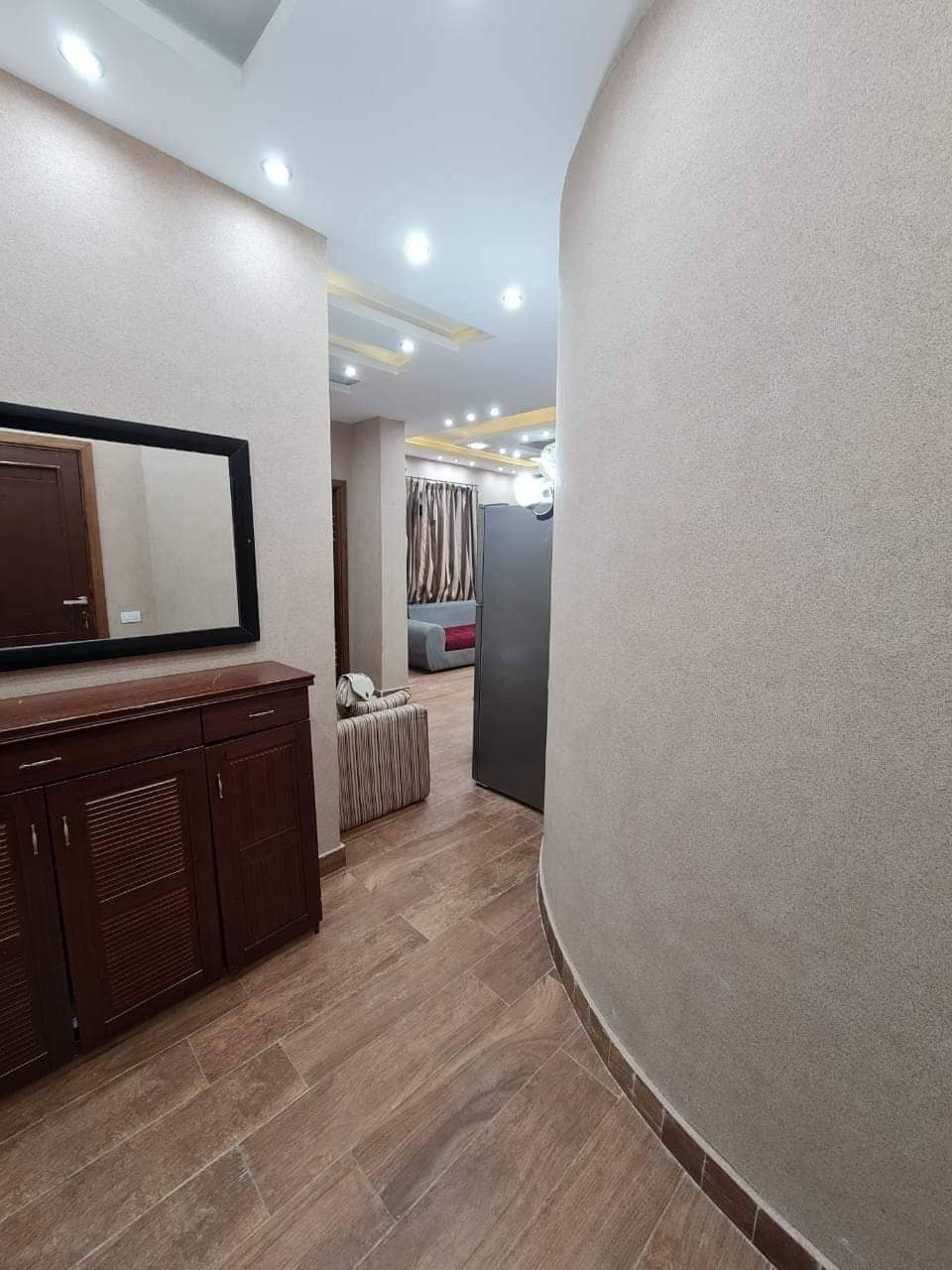 A hallway is shown featuring a wooden cabinet with drawers and a matching mirror. Soft lighting from recessed ceiling fixtures casts a warm glow on the cream-colored walls. The view ahead opens to a spacious living area, adorned with neutral tones and textured curtains.