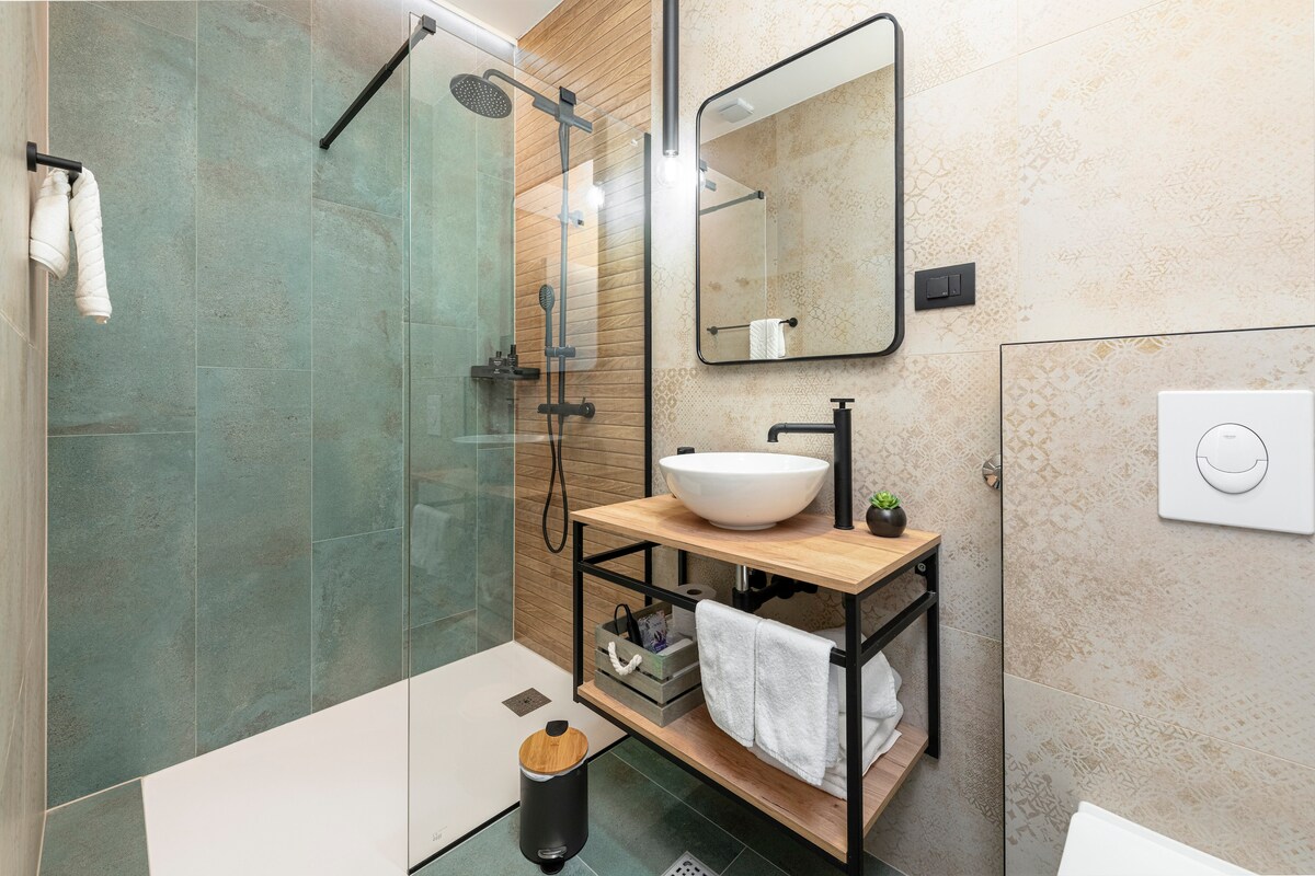 An elegant bathroom is displayed, featuring a walk-in shower enclosed by glass panels. A round sink rests on a wooden vanity with organized toiletries beneath. Soft lighting illuminates the space, and textured tiles in muted tones create a calming atmosphere.