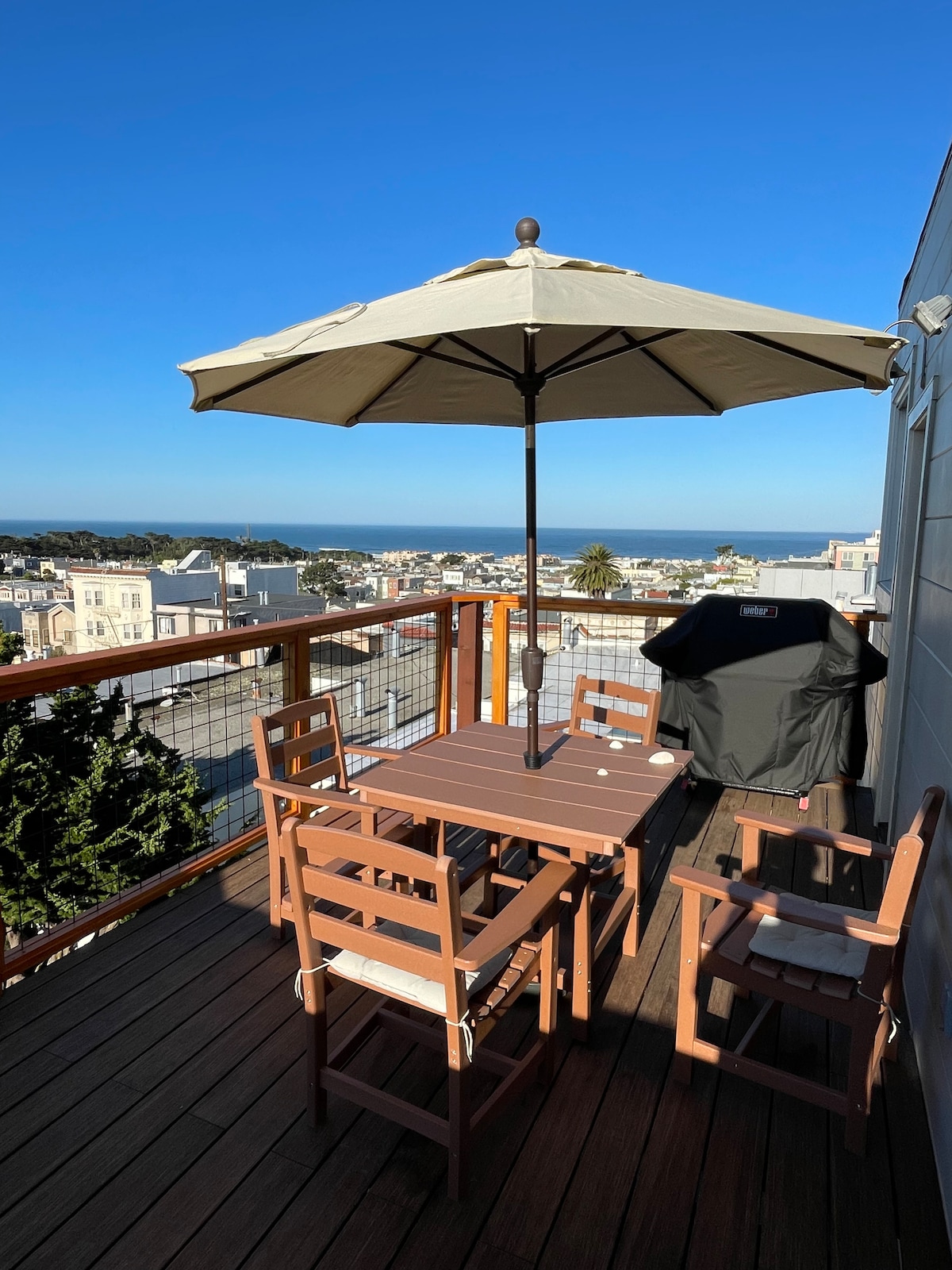 A private deck is presented, featuring a wooden table and four chairs under a large umbrella. A grill is positioned nearby, with views extending towards the ocean in the background and surrounding residential areas. The deck surface is made of wooden planks.