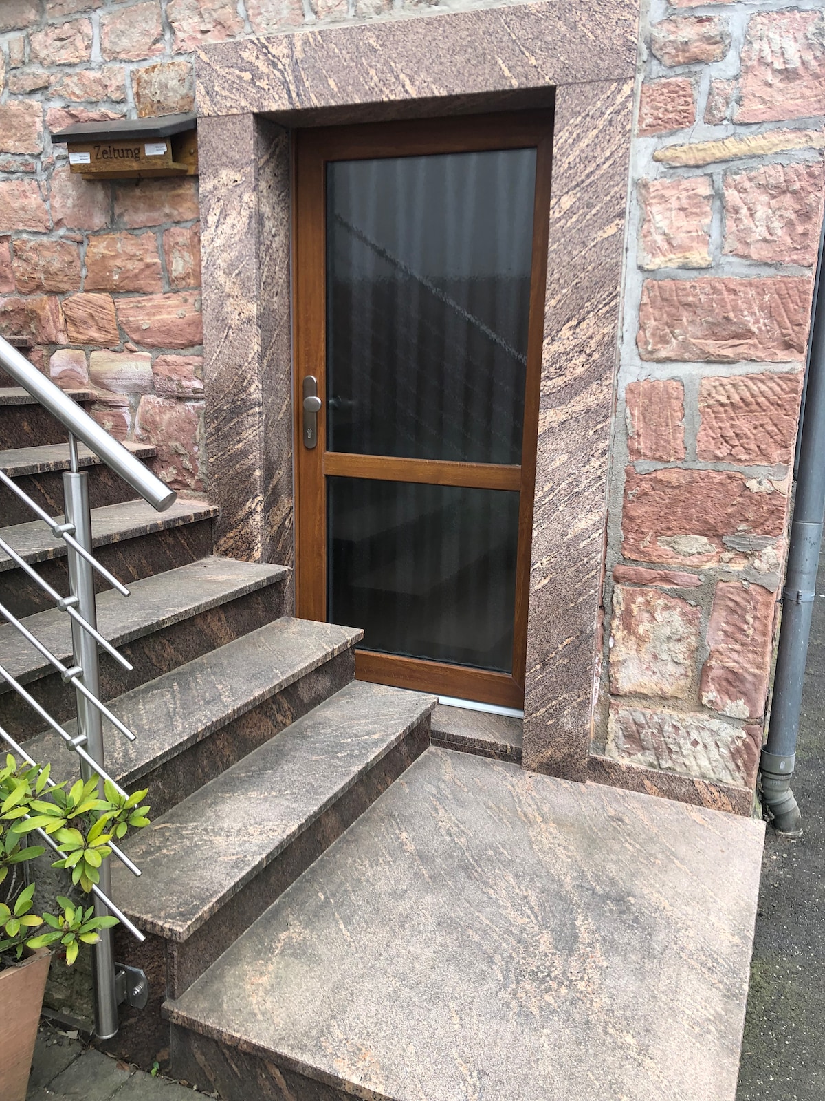 A separate entrance is visible, featuring a wooden door with glass panels. Stone steps lead up to the entrance, complemented by a textured stone wall. A small green plant is positioned near the base of the steps, enhancing the welcoming appearance of the entrance.