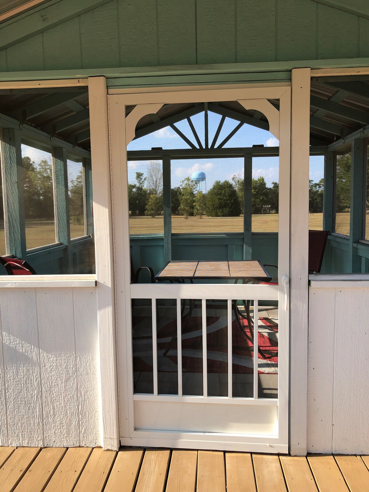 A screened-in porch features a white door with an arched design, leading to a view of an open lawn. A wooden table is positioned inside, surrounded by two black chairs. Natural light filters through the screens, creating a bright and airy space.