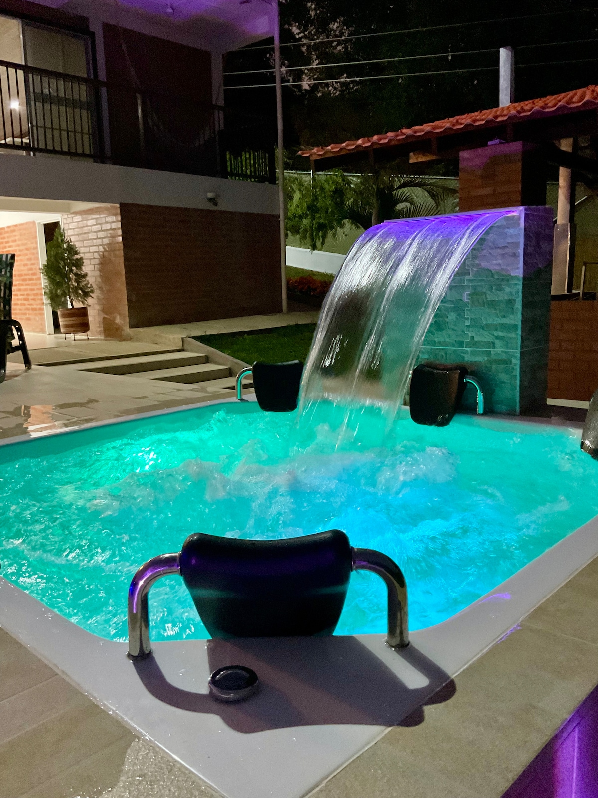 A relaxing jacuzzi is shown, featuring illuminated water in shades of blue and green. A cascading waterfall pours into the jacuzzi, enhancing the tranquil ambiance. The surrounding area includes a paved space and lush greenery, creating a serene outdoor setting.