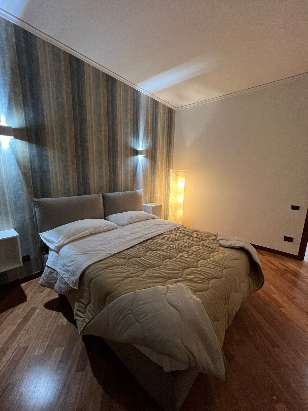 A welcoming bedroom features a spacious bed with a neutral-toned quilt and multiple pillows. Soft lighting from wall sconces and a standing lamp creates a warm ambiance. The striped accent wall adds subtle texture, complemented by wooden flooring.