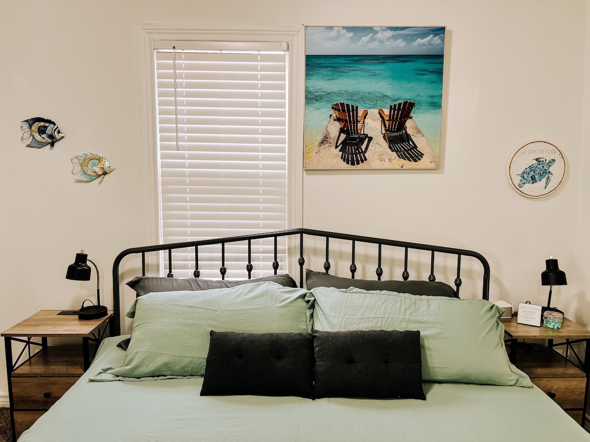 The sleeping area features a king-size bed dressed in light green linens and adorned with black pillows. Adjacent night tables hold stylish lamps. A serene beach-themed artwork enhances the wall above, with a window covered by white blinds allowing natural light.