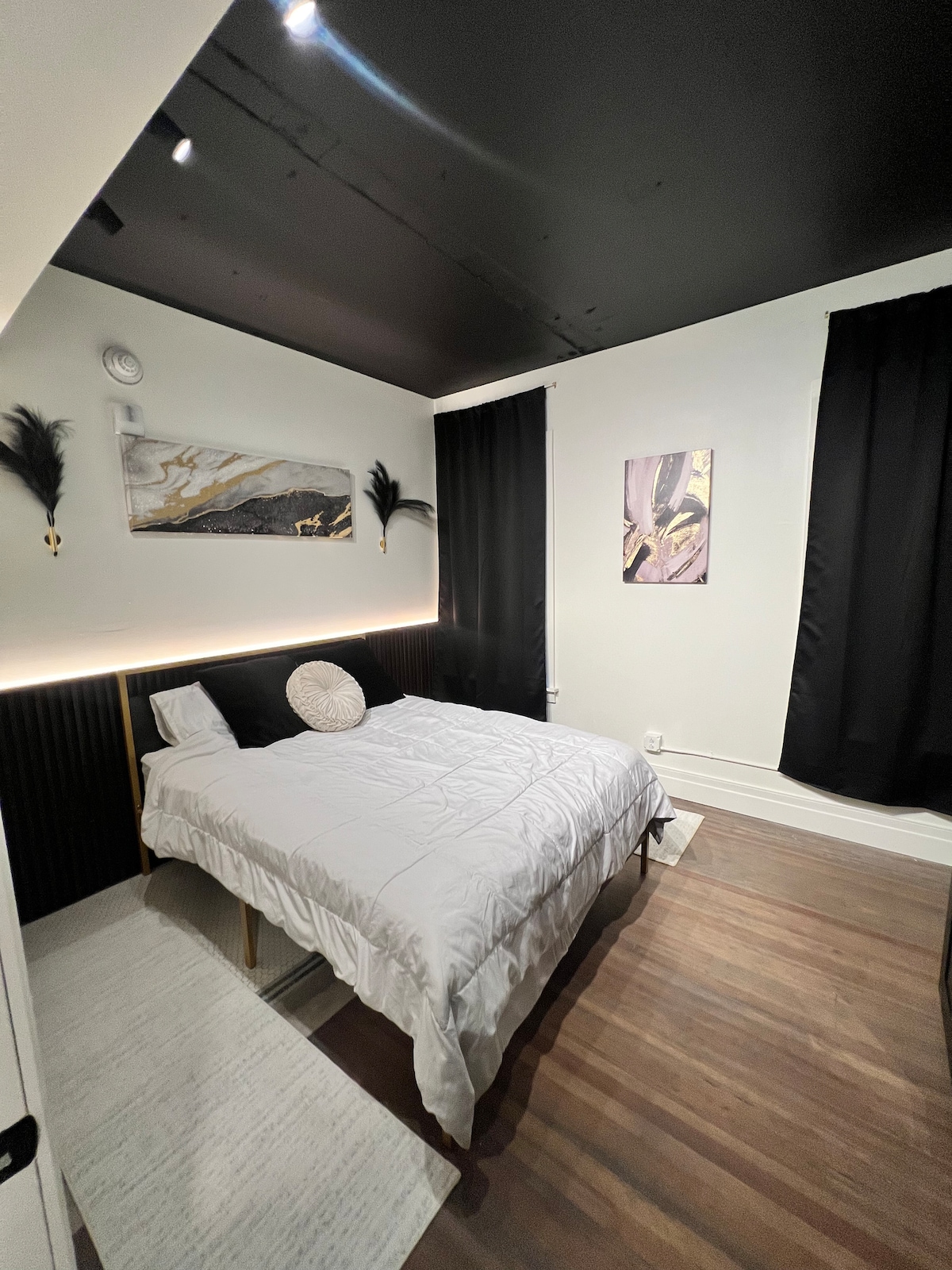 A cozy bedroom is designed with a spacious bed featuring a light comforter. Dark curtains frame the windows, enhancing privacy. A piece of wall art adds a touch of color, while minimalistic decor elements complete the inviting ambiance in the room.