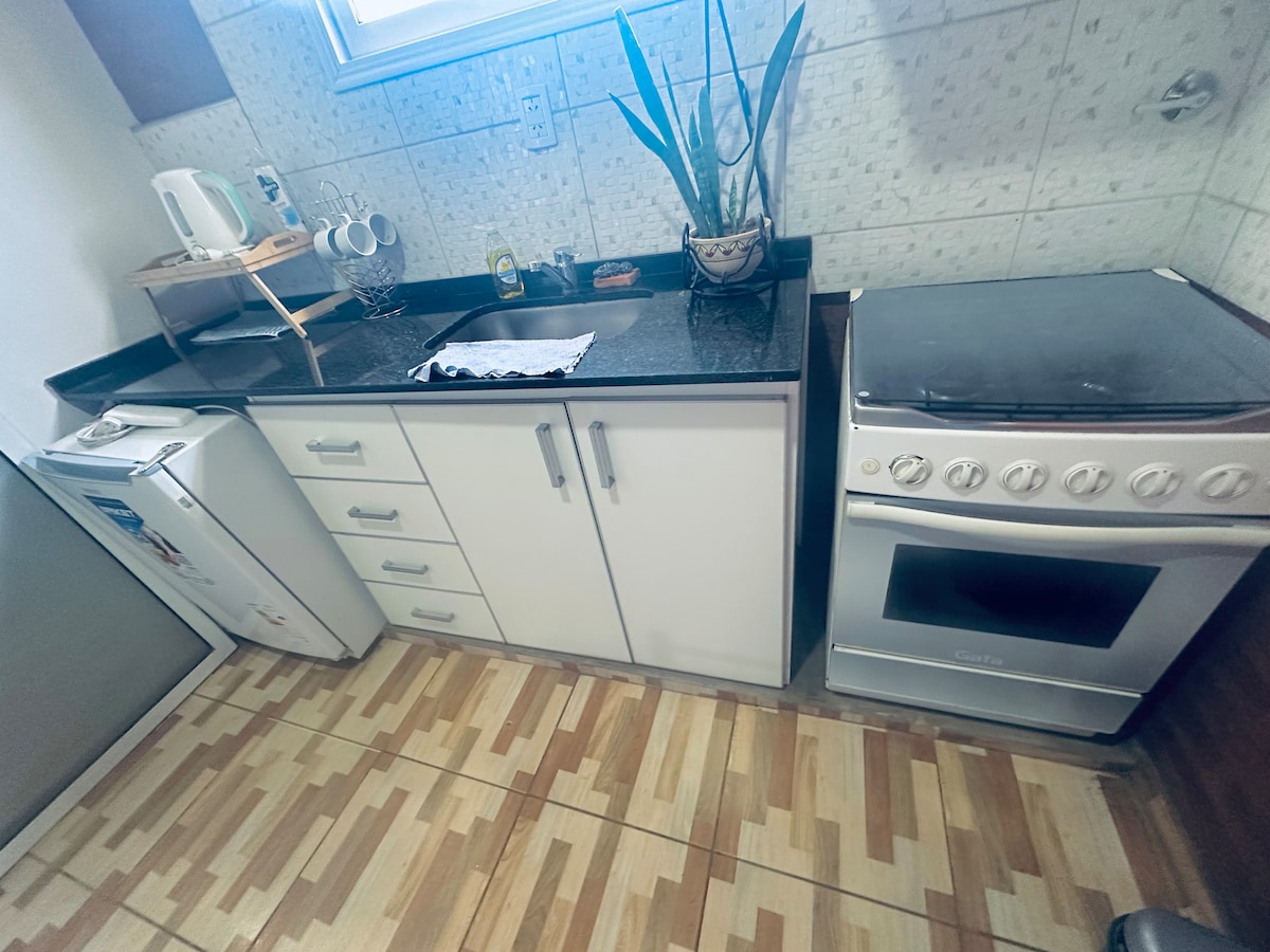 A compact kitchen is equipped with a refrigerator, oven, and a sink with a countertop. Various utensils and appliances, including an electric kettle, are visible. The floor features a wooden pattern, enhancing the kitchen's cozy ambiance.