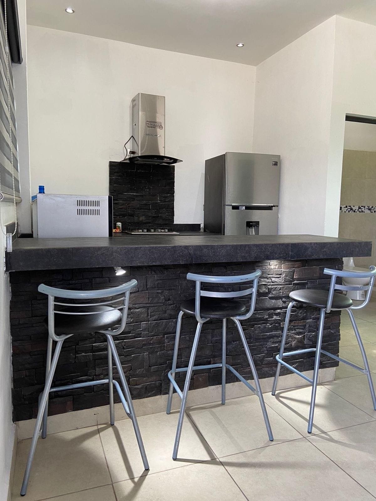 A modern kitchen area is highlighted by a sleek black stone countertop, featuring three high stools for seating. Stainless steel appliances, including a refrigerator and a hooded stove, are positioned against a white wall, contributing to the functional and inviting design.