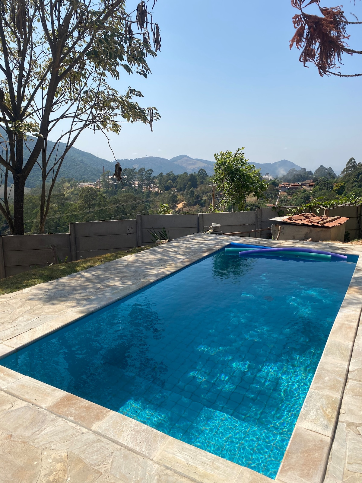 A rectangular swimming pool is surrounded by a paved deck and green grass. The pool's clear blue water reflects the sky, while distant hills and trees create a serene backdrop. A few scattered plants can be seen near the edge of the pool.