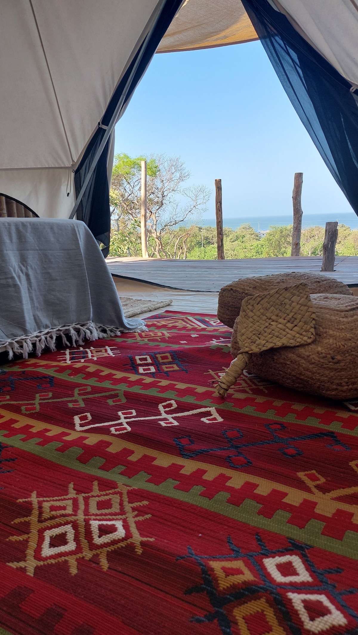 A round tent interior features a colorful woven rug with geometric patterns. A plush seating area is arranged with decorative cushions. The entrance opens to a view of lush greenery and the Caribbean Sea in the distance, allowing natural light to fill the space.