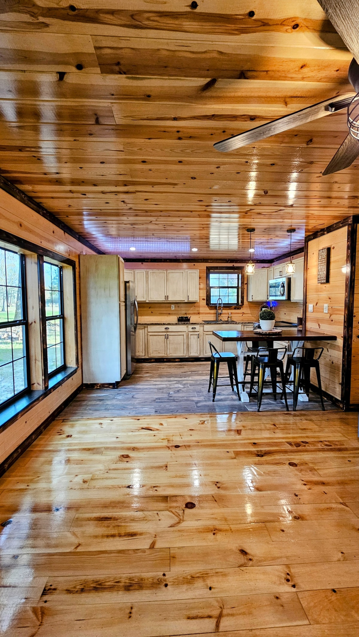 An open interior space showcases a wooden ceiling and polished floors. Large windows provide natural light, highlighting a dining table with chairs and a kitchen area equipped with cabinetry and appliances. The setting conveys a warm and inviting environment, perfect for relaxation.