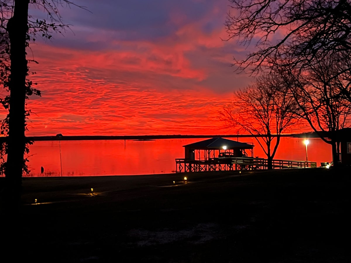 A vibrant sunset is captured over the lake, casting shades of red and orange across the sky. A boat house is silhouetted against the water, with lights illuminating the surrounding area, and trees provide a natural frame along the shoreline.