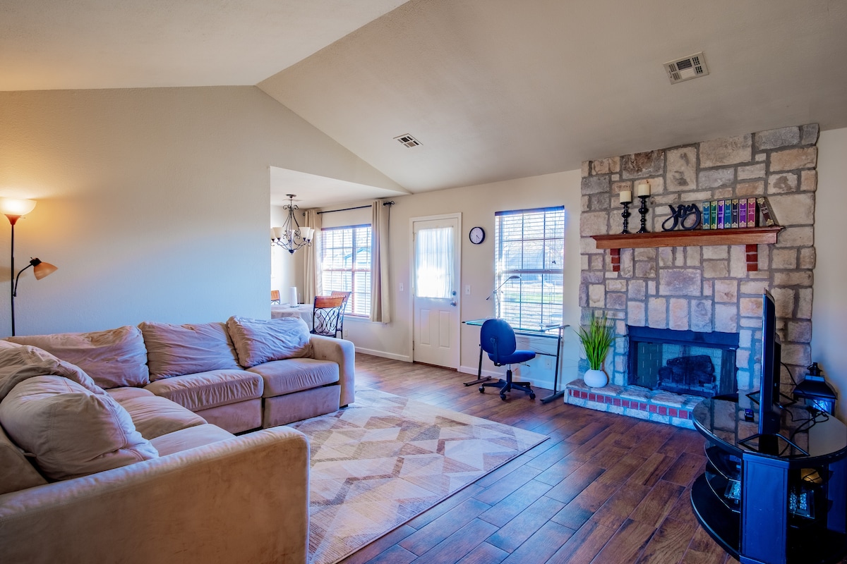 A spacious living area is showcased with a large, comfortable sectional sofa and a stone fireplace. Windows allow natural light to fill the room, creating an inviting atmosphere. A flat-screen TV is positioned near the fireplace, and a desk is situated in the corner.