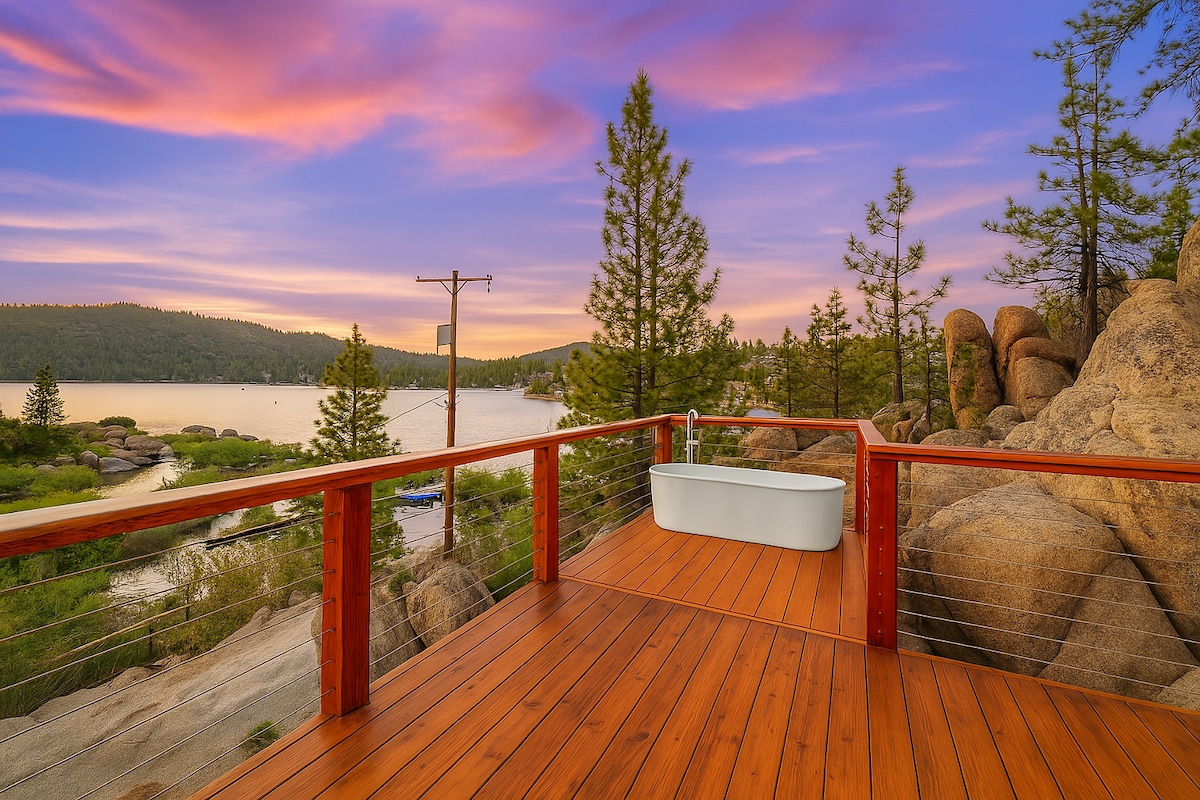 A spacious wooden deck features an outdoor soaking tub against a backdrop of stunning sunset colors. Towering trees and boulders frame the serene lake view, creating a tranquil setting. The railing surrounding the deck enhances safety while providing unobstructed access to the natural beauty.
