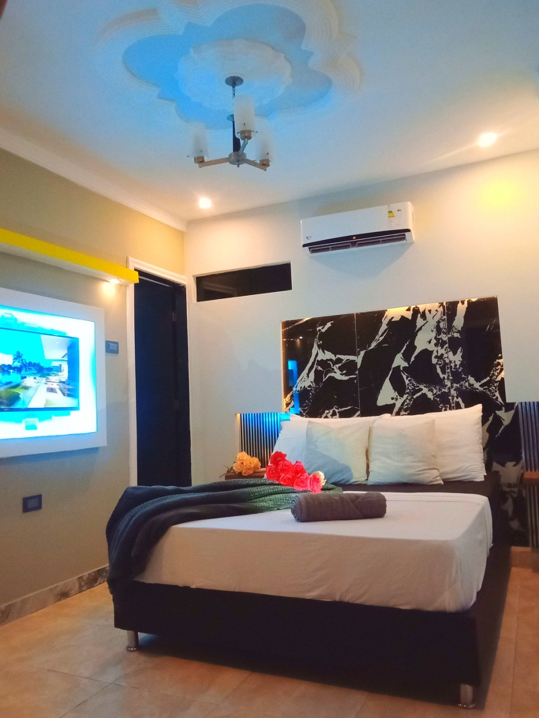 A contemporary sleeping area is highlighted by a stylish headboard and soft bedding. A Smart TV is mounted on the wall, and a floral decoration adds a touch of color. Soft lighting from a ceiling fixture creates a warm ambiance.