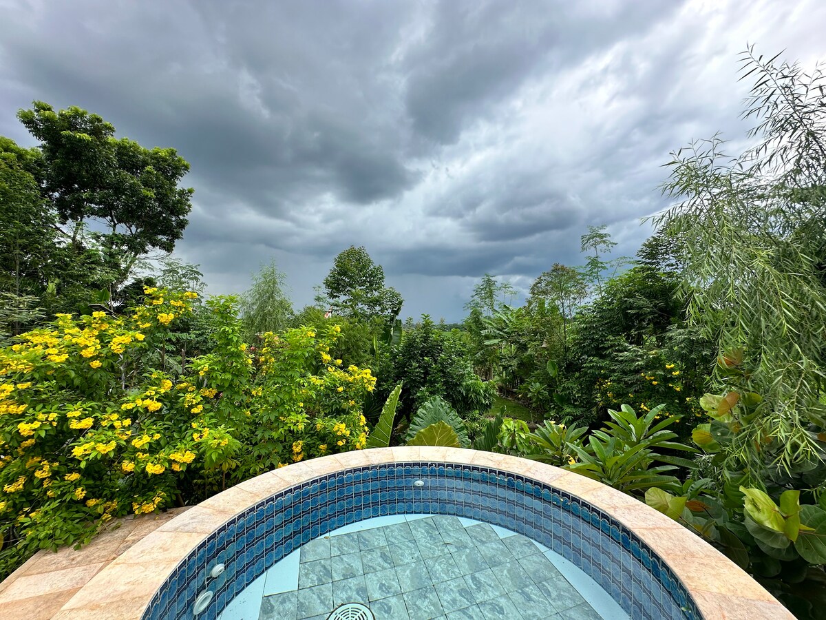 A private jacuzzi is set against a backdrop of lush greenery and vibrant yellow flowers. Dark storm clouds loom above, creating a contrast with the surrounding nature. The jacuzzi features blue tiles that reflect the sky, offering a serene outdoor experience.