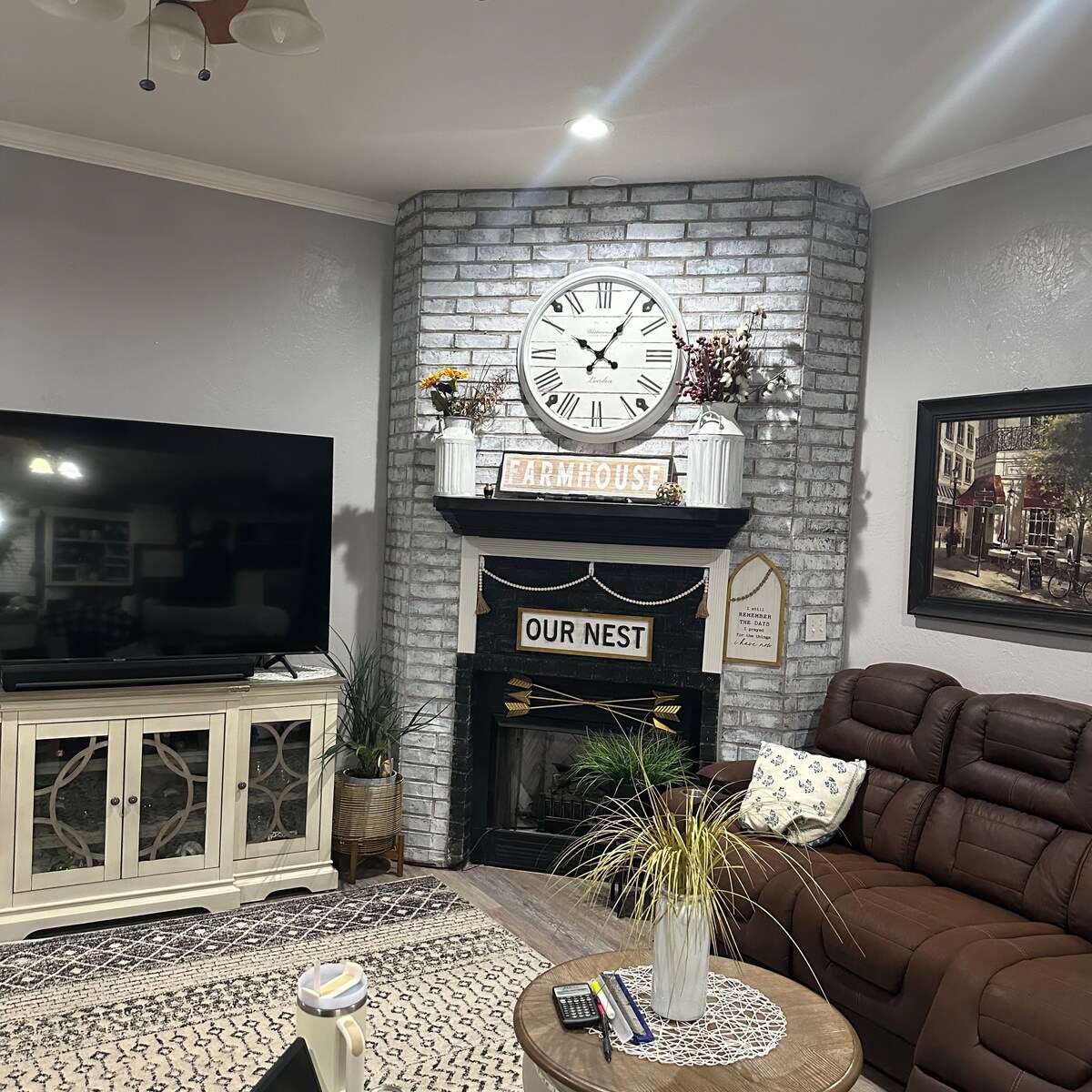 A cozy living room features a large dark brown sectional sofa and a rustic coffee table. A wall-mounted television is positioned across from the seating area. A decorative brick fireplace is framed by a white mantle adorned with plants and a clock, complemented by wall art.