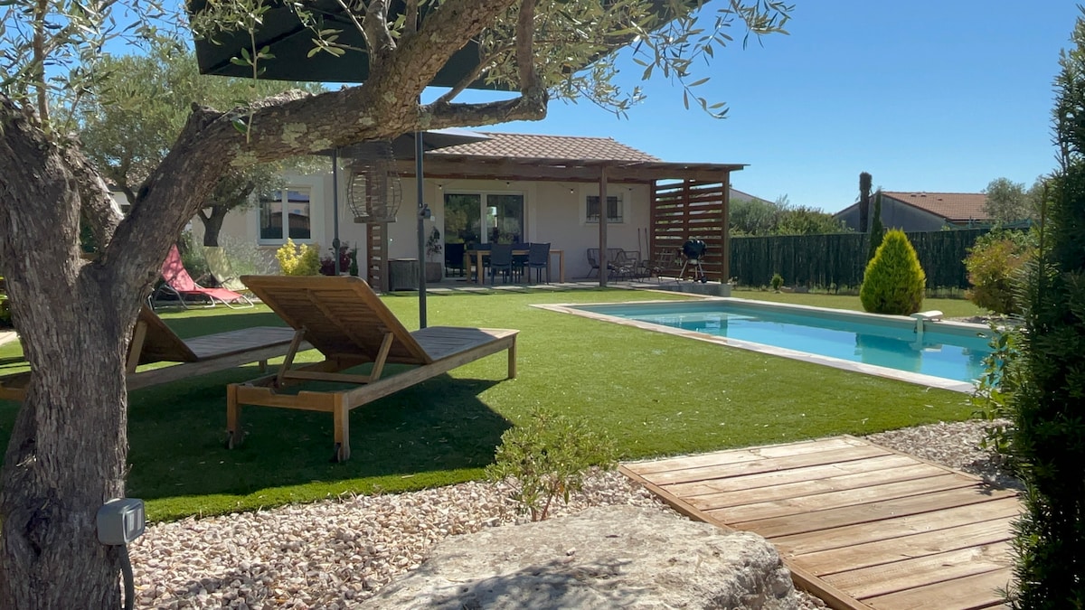 A serene outdoor space is featured, showcasing a well-kept lawn with sun loungers positioned for relaxation. A sparkling pool reflects the clear blue sky, while a shaded area with a dining table invites gatherings. A tree offers natural shade, enhancing the tranquil setting.