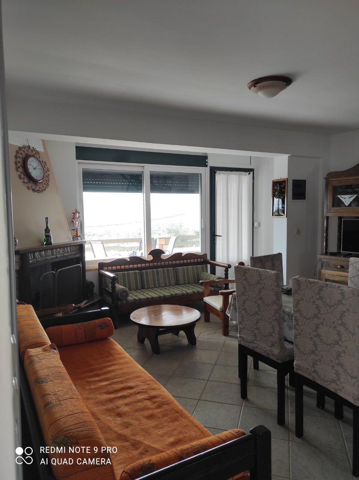 A living area is featured with two sofas positioned around a circular wooden coffee table. A dining table with upholstered chairs is visible. Large windows provide natural light and views, while a decorative clock and a small shelf add character to the space.