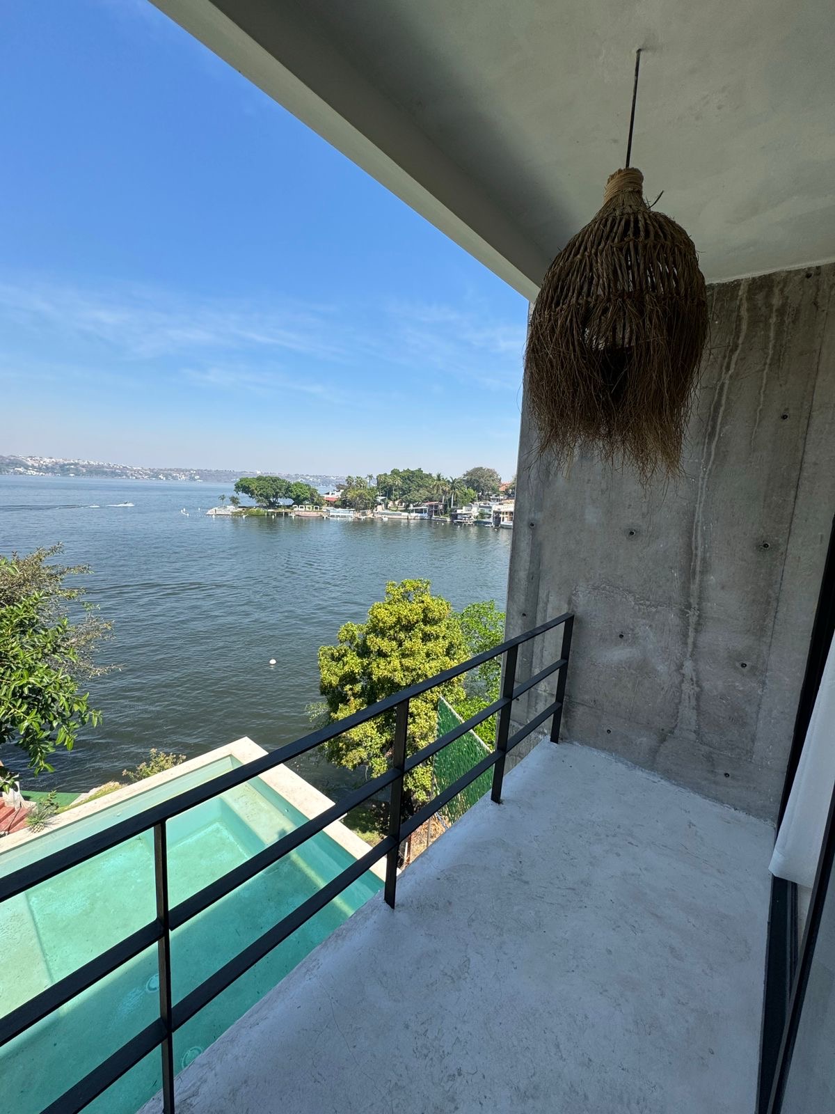 A balcony offers views of the shimmering lake, framed by lush greenery. A hanging woven light fixture adds a natural touch above the concrete floor, creating a serene space to enjoy the surroundings. The clear blue sky enhances the tranquil atmosphere.