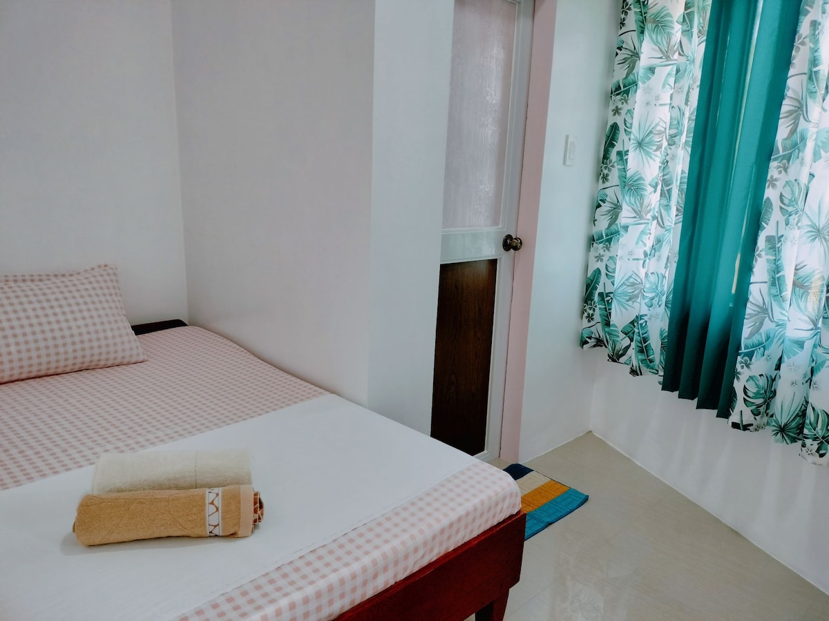 A cozy room features a double bed with a checkered bedspread and neatly folded towels. Natural light streams in through the window, framed by green curtains with a floral pattern. The floor is decorated with a small, colorful rug.