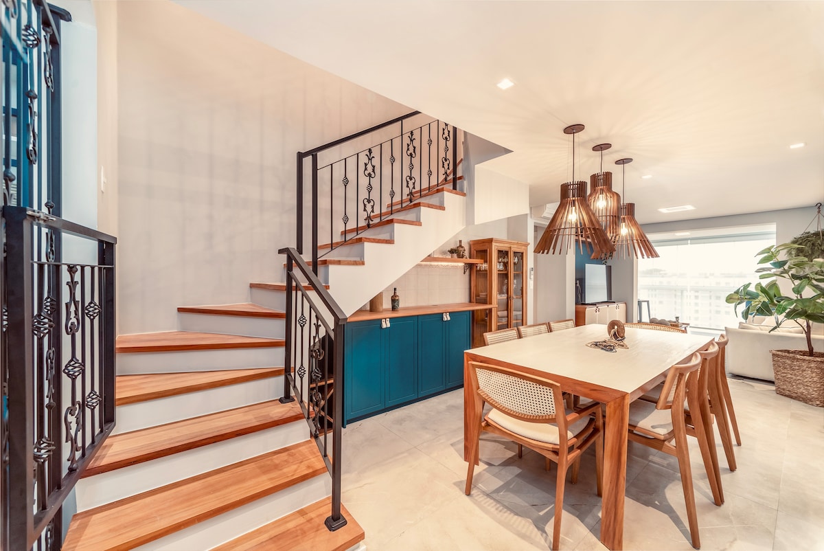 A modern dining area is featured with a large wooden table surrounded by light-colored chairs. A stylish light fixture hangs above, and a staircase with wooden steps and wrought iron railings leads to an upper level. Natural light fills the space from a nearby window.