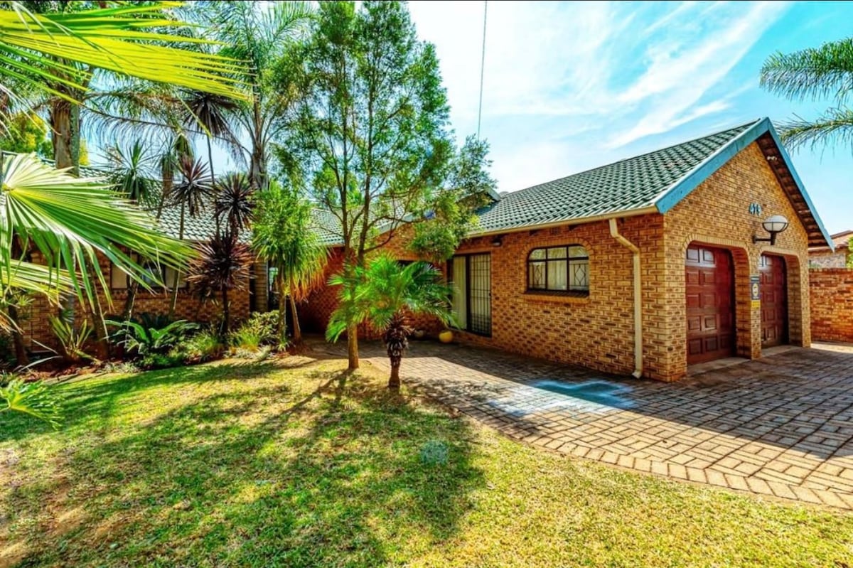 The exterior of the home features a welcoming brick façade surrounded by lush greenery, including palm trees and shrubs. A private driveway leads to the double garage, and a well-maintained lawn adds to the inviting atmosphere.
