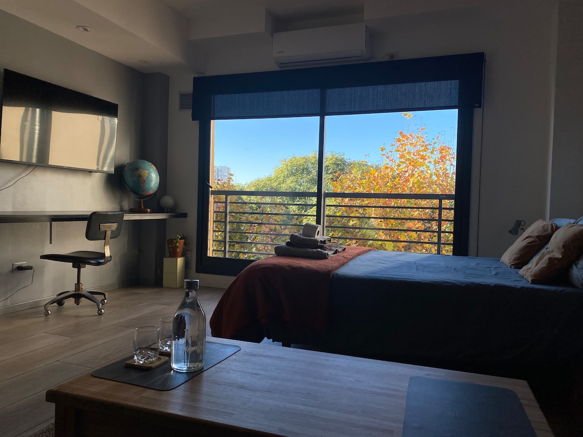 A cozy living space features a spacious bed dressed in warm tones, with a desk and chair positioned nearby. A window offers a clear view of greenery and trees outside, while natural light fills the room. A small table is set with glassware.