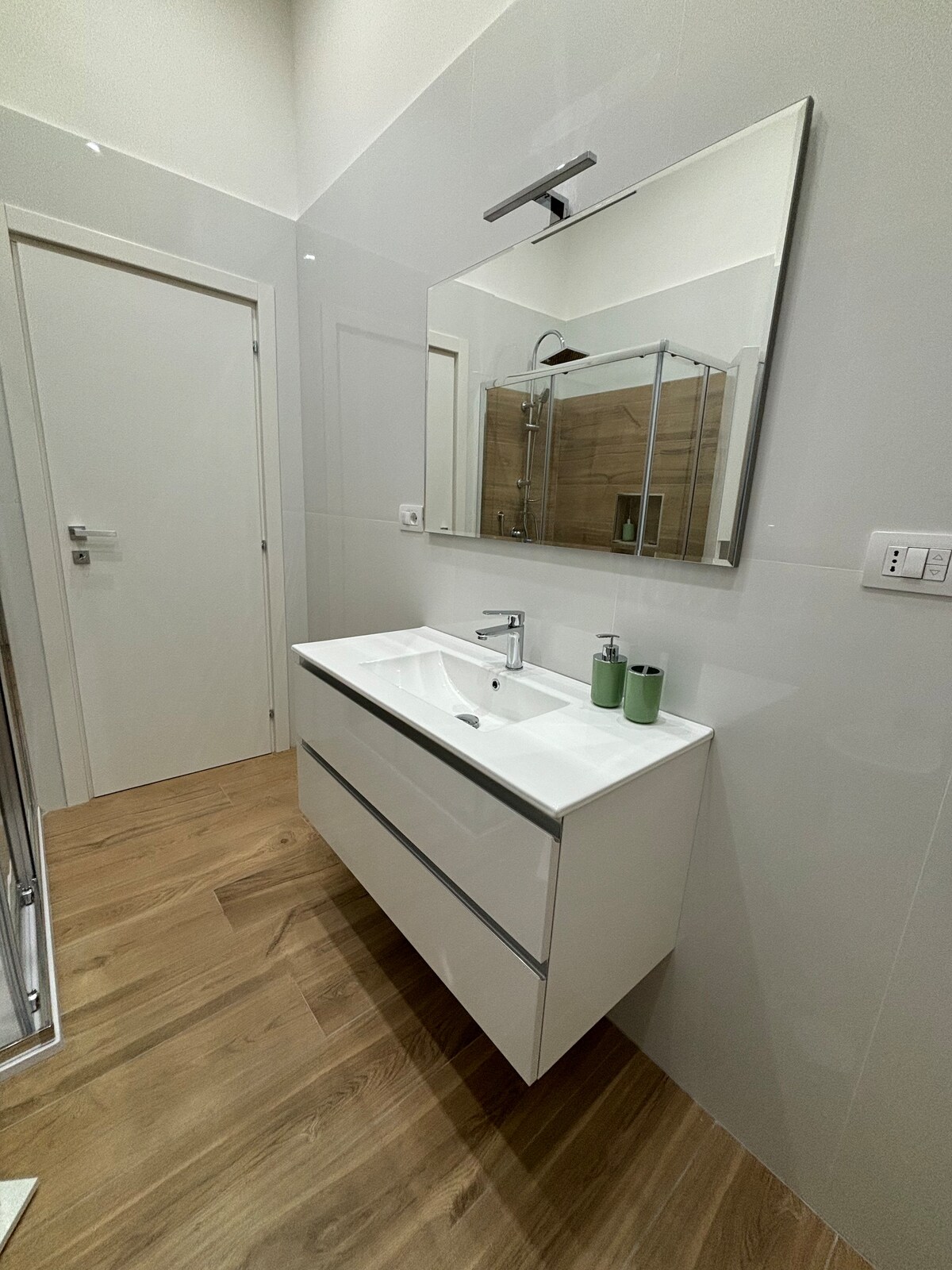 A modern bathroom features a sleek white vanity unit with two drawers and a porcelain sink. A large mirror is mounted above the sink, reflecting the room's clean lines. Natural wood accents can be seen on the floor and in the shower area beyond.