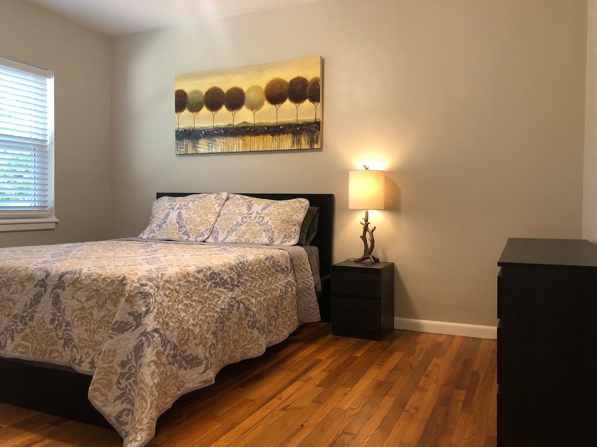 A well-appointed bedroom features a spacious bed covered with a patterned quilt, complemented by a bedside table with a lamp. Natural light flows through a window with blinds, and a dresser is positioned nearby on polished hardwood flooring, creating a calm and functional space.