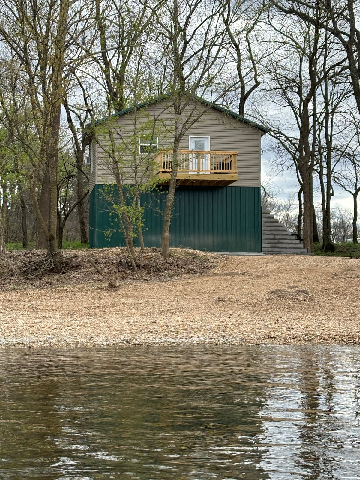 A cabin elevated on a green metal base overlooks a creek, surrounded by trees. Sturdy steps lead up to a small balcony with wooden railings. The shoreline is lined with smooth gravel and boulders, reflecting a calm water surface below.