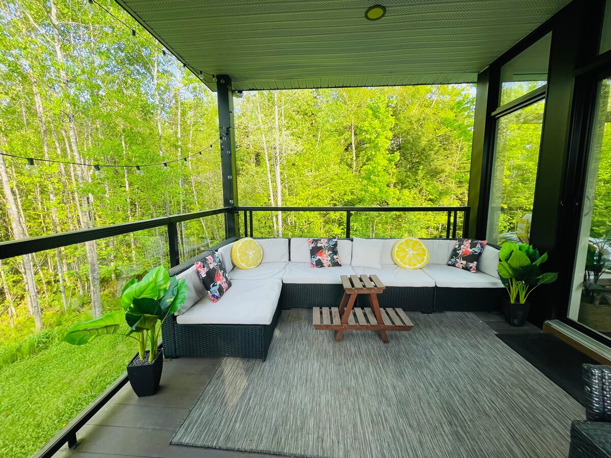 A covered terrace features a spacious seating area with a sectional sofa adorned with decorative pillows. A small coffee table sits in the center, surrounded by lush greenery visible through large windows. Potted plants enhance the inviting atmosphere.