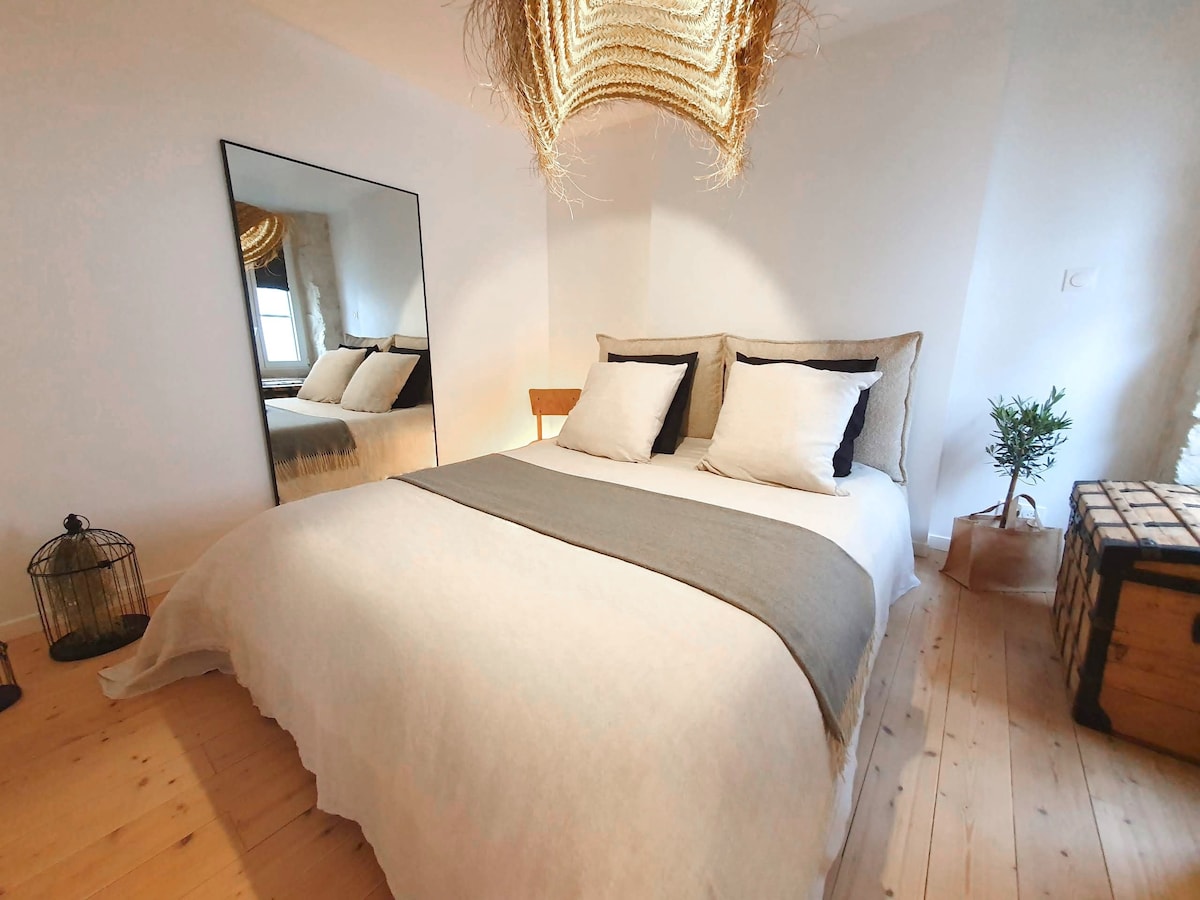 The serene bedroom features a spacious bed with light-colored linens and four pillows, complemented by a soft throw. A mirrored wardrobe reflects the room, while a small plant adds a touch of greenery. Natural light fills the space, accentuated by a woven light fixture.