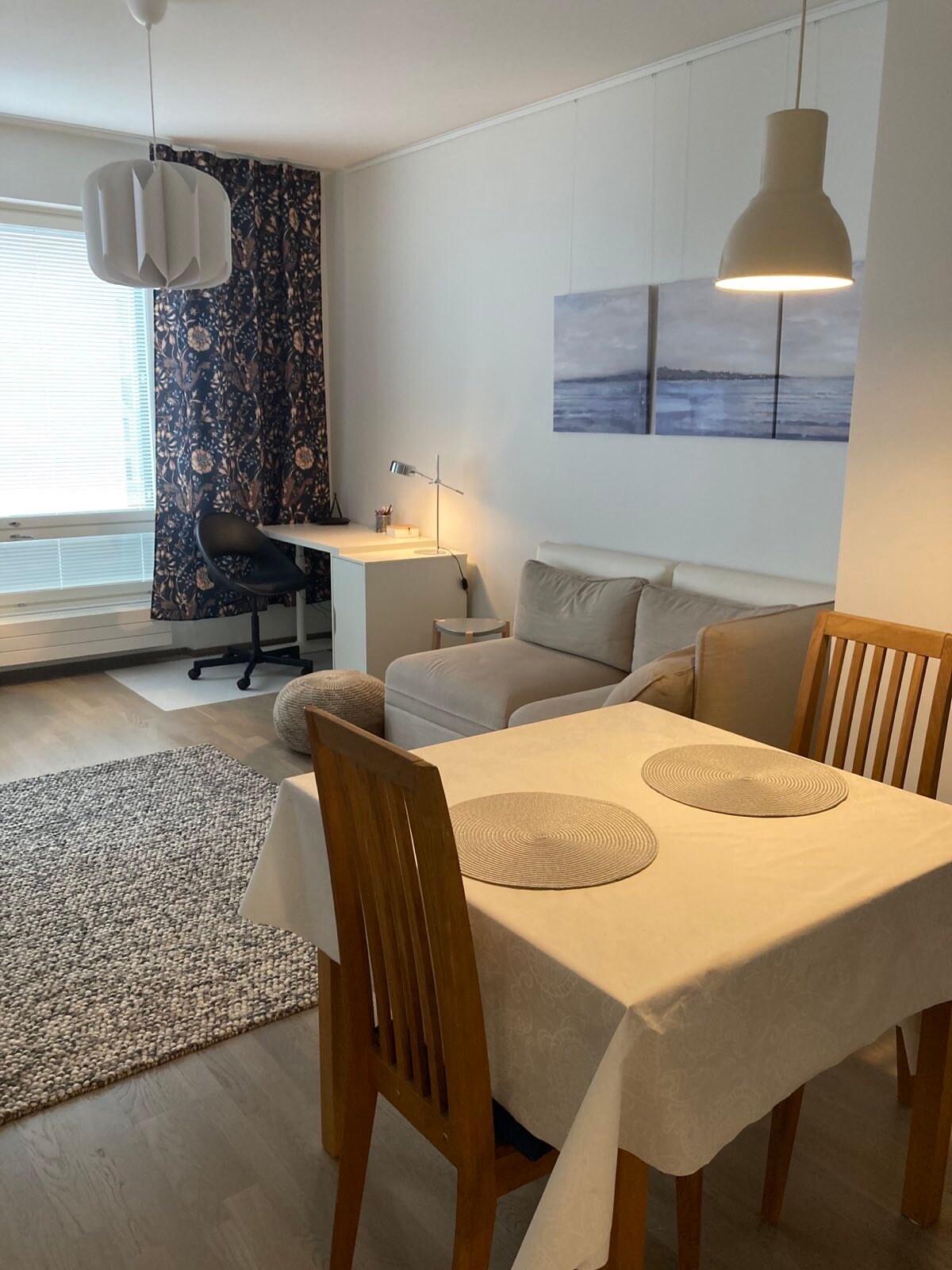 A modern living space features a light-colored sofa and a small desk with a task lamp. A dining table with two place settings is set nearby. Large windows dressed with patterned curtains allow natural light to fill the room, while a textured area rug adds warmth.