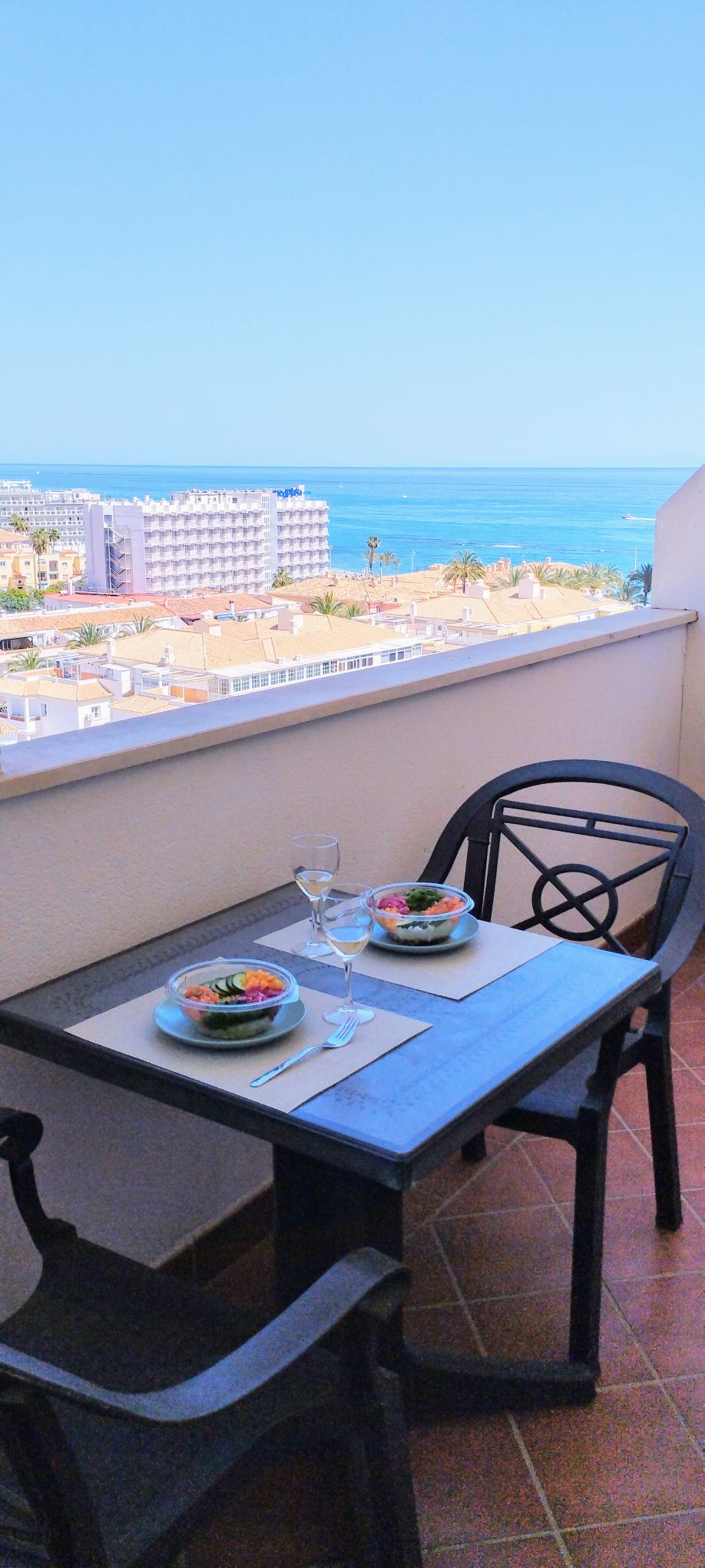 A balcony table set for two features plates with salads and glasses. Expansive views of the coastline and sea are visible in the distance, framed by the warm light of a clear sky. The table is positioned against a neutral-colored wall.