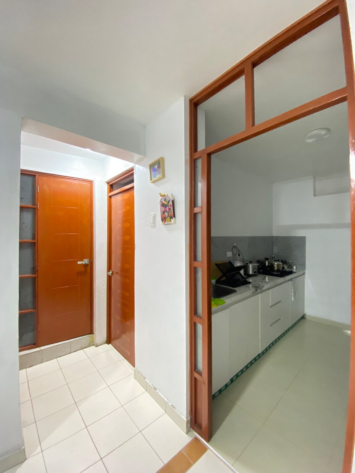 A well-lit corridor leads to a modern kitchen area, equipped with essential appliances. Two wooden doors are visible on the left, providing access to private rooms. The kitchen features white cabinetry and a tiled backsplash, complemented by ample counter space.