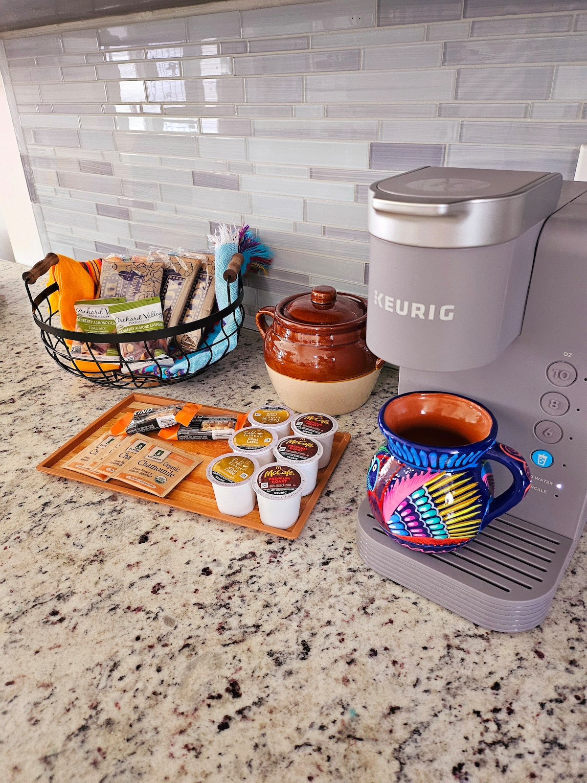 A countertop showcases a Keurig coffee maker alongside a variety of coffee pods arranged on a wooden tray. A colorful mug rests beside the machine, while a basket contains assorted snacks and a ceramic pot, contributing to a homely kitchen ambiance.